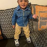 toddler, child, denim_jacket, khaki_pants, sneakers, necklace, couch, cushions, patterned, indoor, smiling, standing, person, young_child, casual_clothing, cute, portrait, home, fashion, happy