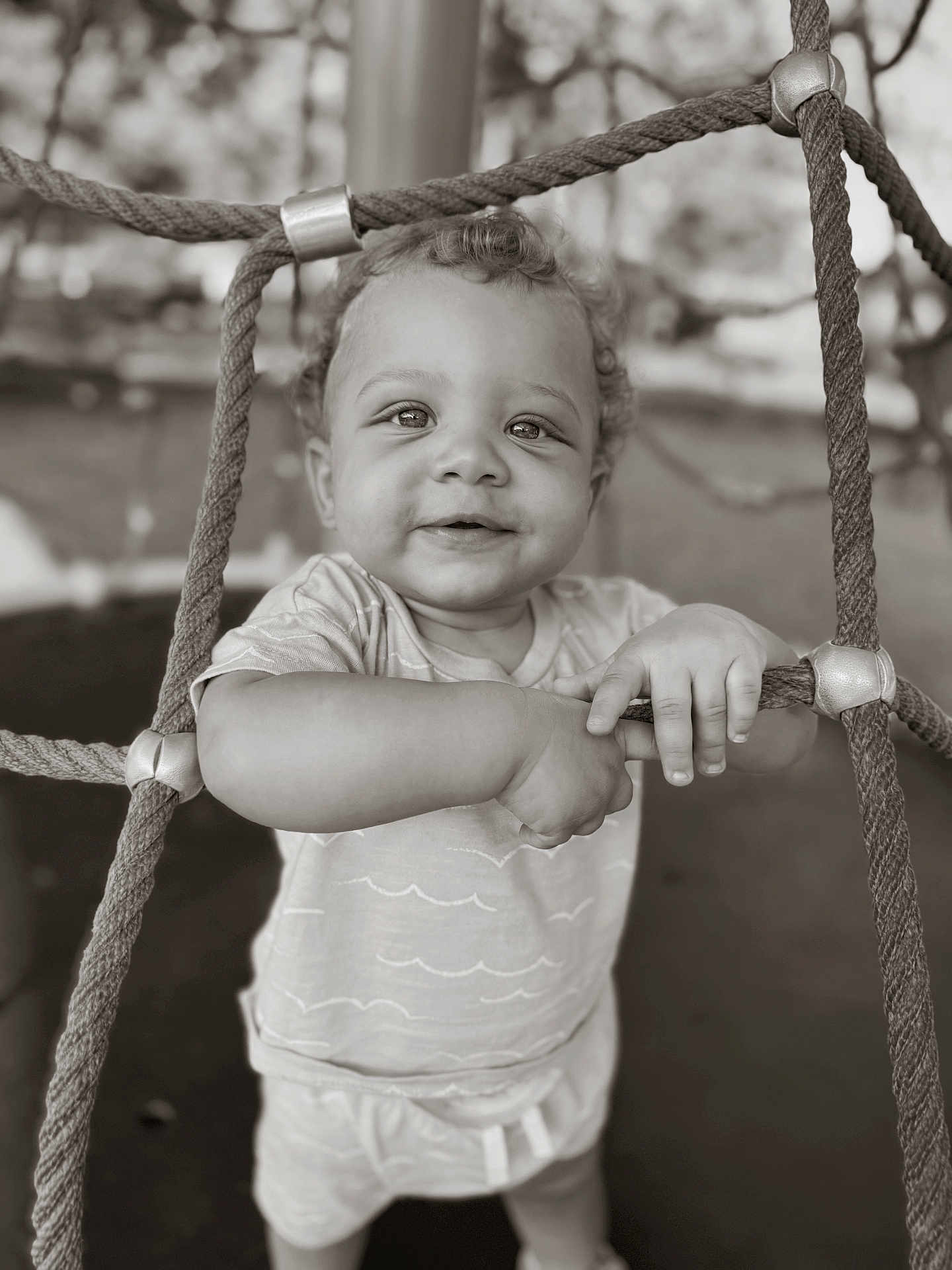 Adrian is registered to the contest to win money with this photo: toddler, child, playground, rope, net, smile, curly_hair, monochrome, outdoor, baby, grip, cute, person, shirt, shorts, expression, young_child, play, fun, daytime