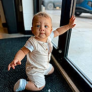 Adrian is registered to the contest to win money with this photo: baby, child, blue_eyes, romper, indoor, kneeling, carpet, glass_door, hand, curious, blond_hair, footwear, shoes, window, natural_light, expression, person, floor, background, daylight