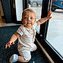 baby, child, blue_eyes, romper, indoor, kneeling, carpet, glass_door, hand, curious, blond_hair, footwear, shoes, window, natural_light, expression, person, floor, background, daylight