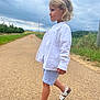 child, blonde_hair, white_shirt, striped_shorts, sandals, walking, road, country_road, outdoor, grass, cloudy_sky, rural, nature, side_view, person, summer, casual_clothing, young_child, portrait, daytime