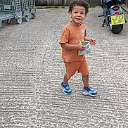 Victor is registered to the contest to win money with this photo: child, boy, orange_clothing, sneakers, snack, concrete, outdoor, fence, wooden_fence, motorcycle, metal_carts, smiling, walking, daylight, casual, urban, young_child, happy, person, playful