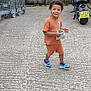 child, boy, orange_clothing, sneakers, snack, concrete, outdoor, fence, wooden_fence, motorcycle, metal_carts, smiling, walking, daylight, casual, urban, young_child, happy, person, playful