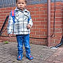 Victor is registered to the contest to win money with this photo: child, boy, standing, brick_wall, brick_pavement, backpack, plaid_jacket, blue_jeans, shoes, outdoor, window, pipe, curb, sidewalk, serious_expression, person, clothing, casual, daylight, urban
