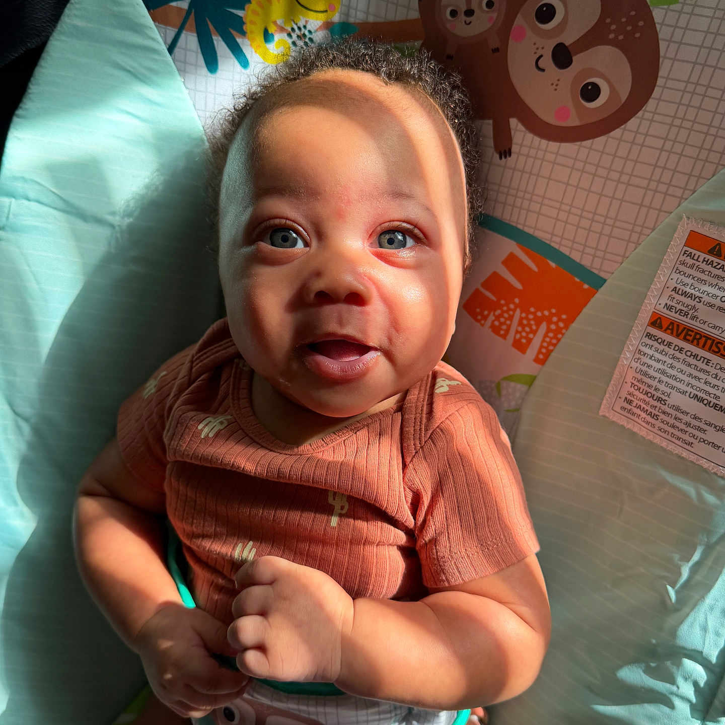 Torran is registered to the contest to win money with this photo: baby, infant, child, play_mat, sunlight, shadow, face, blue_eyes, curly_hair, rust_colored_shirt, bib, sloth, cartoon, animal_graphic, smile, hands, indoor, colorful, cute, portrait