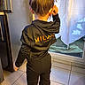 child, back_view, black_clothing, indoor, tiled_floor, curtain, glass_door, hair_bun, standing, person, kitchen, natural_light, floor, scratching_ear, casual_wear, home, young_child, text_on_clothing, long_sleeves, pants