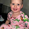 child, toddler, smiling, pajamas, indoor, blue_eyes, hair_bun, cute, happy, sitting, cartoon_print, pink_clothing, foot, person, young_child, face, skin, cheeks, bright_eyes, cozy