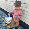child, boy, red_shirt, tan_pants, black_shoes, blue_egg, basket, gravel, house_wall, outdoor, smiling, curly_hair, playful, spring, daylight, casual_clothing, holding, holiday, fun, exploring