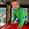 child, playground, climbing, green_jacket, hood, red_play_structure, mesh_grid, wooden_frame, indoor_play_area, smiling, blue_eyes, patterned_pants, hand, crouching, play, young_child, casual_clothing, bright_colors, fun, active
