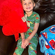 Eli is registered to the contest to win money with this photo: child, smiling, dinosaur_pajamas, red_pillow, blue_blanket, couch, leather, indoor, happy, sitting, plush, cozy, tag, young_child, casual, relaxed, portrait, cute, colorful, home