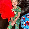 child, smiling, dinosaur_pajamas, red_pillow, blue_blanket, couch, leather, indoor, happy, sitting, plush, cozy, tag, young_child, casual, relaxed, portrait, cute, colorful, home