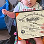baby, child, diploma, certificate, bow, plaid_pants, person, indoor, yellow_wall, blue_bag, sitting, clothing, hand, foot, face, smiling, portrait, holding, toddler, proud