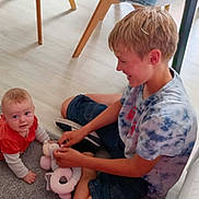 Enzo a rejoint le concours — aidez-le/la à gagner de superbes lots ! baby, child, indoor, floor, toy, stuffed_animal, carpet, sitting, playing, smiling, person, shorts, tshirt, wood_floor, furniture, light, happy, candid, family, interaction
