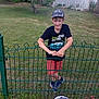 boy, child, smiling, cap, tshirt, shorts, fence, grass, backyard, tree, sports_equipment, badminton, shuttlecock, outdoor, play, summer, recreation, person, casual_wear, happy