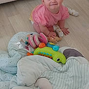 Naomie participe au concours pour gagner de l'argent avec cette photo : baby, blanket, child, colorful, couch, crawling, cute, floor, happy, head, indoor, infant, person, pink_shirt, playful, smiling, sock, soft_toys, toys, wooden_floor