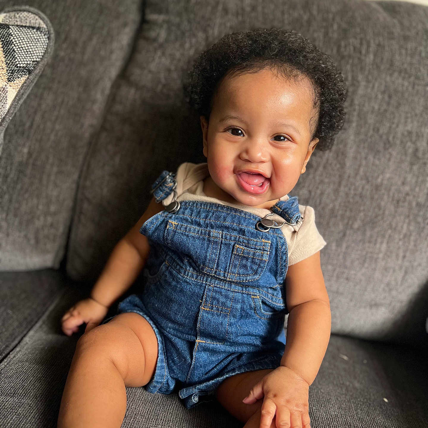 Zadkiel is registered to the contest to win money with this photo: baby, casual_clothing, child, comfortable, couch, curly_hair, cute, denim_overalls, happy, indoor, infant, one_person, person, portrait, sitting, skin, smiling, sock, toddler, young_child