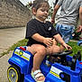 toddler, toy_car, blue, outdoor, child, grass, sidewalk, black_shirt, sandals, steering_wheel, person, summer, playing, vehicle, plastic, fun, kids, casual_clothing, shorts, daytime