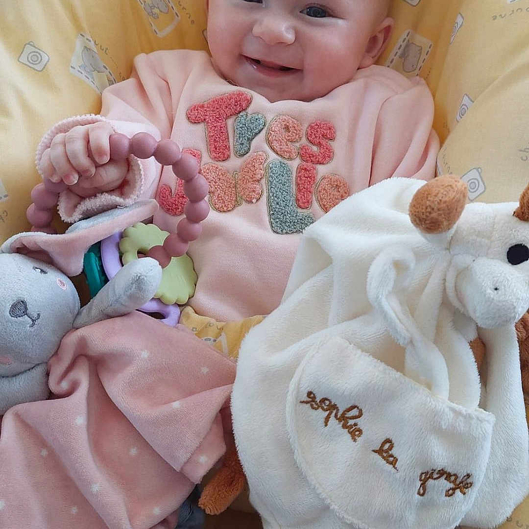 Liv participe au concours pour gagner de l'argent avec cette photo : baby, child, clothing, comfort, cozy, cute, happy, indoors, infant, person, pink_clothing, plush_toy, portrait, sitting, smiling, soft_toys, teething_ring, toy, warm, yellow_cushion