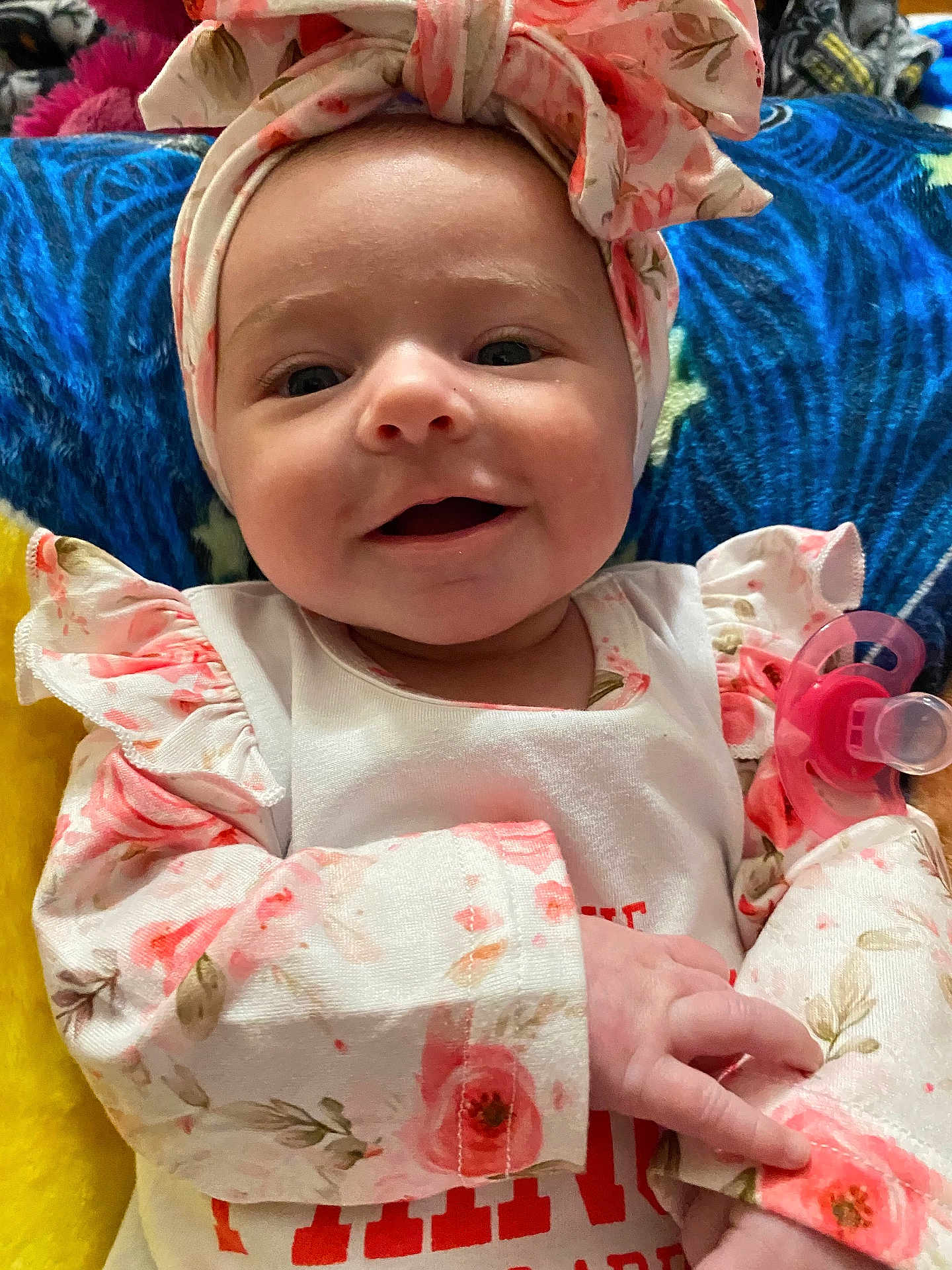 Coraline is registered to the contest to win money with this photo: baby, child, smiling, headband, floral, clothing, pacifier, blanket, cute, infant, portrait, happy, young, lying_down, closeup, person, soft, colorful, indoors, hand