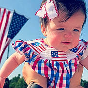 Adalou is registered to the contest to win money with this photo: baby, child, infant, face, person, blue_sky, american_flag, bow, hair_accessory, hand, outdoor, daylight, dress, patterned_clothing, patriotic, red_white_blue, holding, cute, happy, portrait