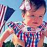 baby, child, infant, face, person, blue_sky, american_flag, bow, hair_accessory, hand, outdoor, daylight, dress, patterned_clothing, patriotic, red_white_blue, holding, cute, happy, portrait