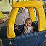 blue, child, cushion, daylight, furniture, happy, outdoor, person, plastic, play, porch, seat, small_vehicle, smiling, steering_wheel, toddler, toy_car, window, wooden_floor, yellow