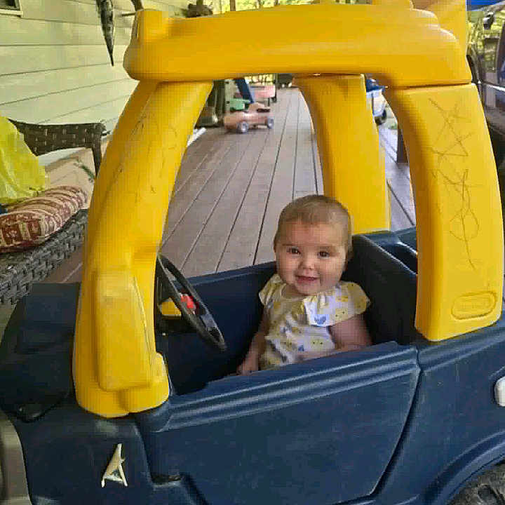 Ryleigh is registered to the contest to win money with this photo: blue, child, cushion, daylight, furniture, happy, outdoor, person, plastic, play, porch, seat, small_vehicle, smiling, steering_wheel, toddler, toy_car, window, wooden_floor, yellow