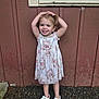 child, girl, smiling, floral_dress, white_sandals, outdoor, gravel_ground, wooden_wall, playful, posing, happy, casual_clothing, daylight, standing, cute, person, portrait, young_child, summer, natural_light