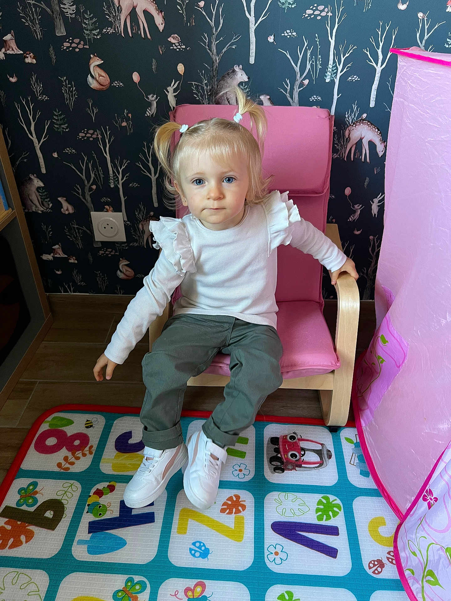 Héloïse a rejoint le concours — aidez-le/la à gagner de superbes lots ! toddler, child, girl, pink_chair, pigtails, white_shirt, green_pants, white_shoes, play_mat, alphabet, colorful, indoor, wood_floor, wallpaper, forest_theme, animals, toy_car, curious, sitting, room