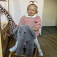 Héloïse a rejoint le concours — aidez-le/la à gagner de superbes lots ! toddler, child, smile, pink_dress, pony_tail, stuffed_elephant, rocking_toy, giraffe_toy, crib, wooden_floor, indoor, playroom, toy, shoes, white_tights, happy, face, person, furniture, home