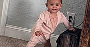 Alora is registered to the contest to win money with this photo: baby, infant, child, standing, pink_onesie, carpet, file_organizer, furniture, wall, power_outlet, smiling, indoor, home, curious, barefoot, person, head, face, hand, support