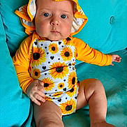 Sabrina is registered to the contest to win money with this photo: baby, child, sunflower, hat, yellow, orange, blue_couch, infant, clothing, portrait, cute, indoors, sitting, one_person, face, skin, legs, hand, foot, expression