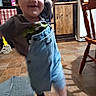 child, toddler, denim_overalls, tshirt, indoor, kitchen, floor, chair, cabinet, mop, rug, smiling, movement, blurry, socks, hat, person, home, playful, young