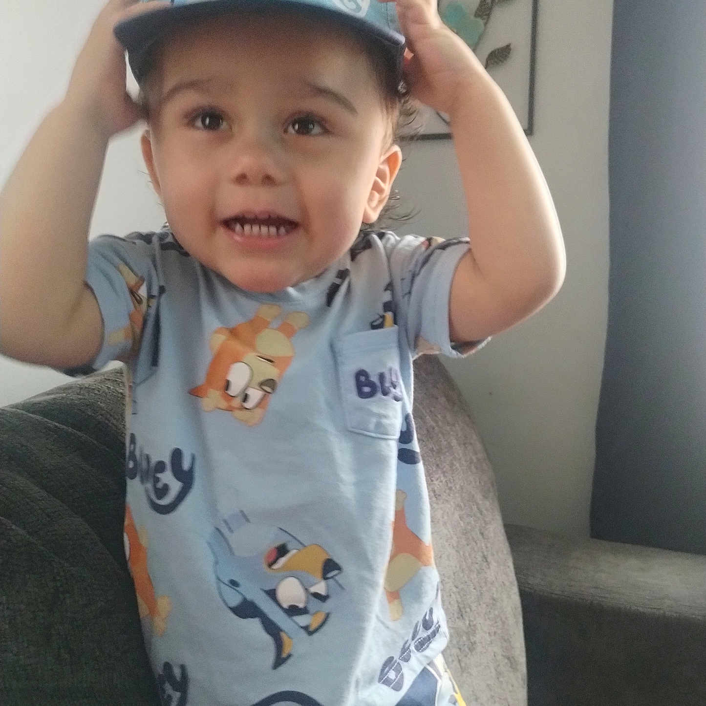 Juilo is registered to the contest to win money with this photo: toddler, child, hat, bluey, shirt, couch, living_room, smiling, happy, person, indoors, furniture, home, decor, wall_art, short_sleeves, gesture, casual_clothing, young_child, cute