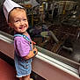 child, curly_hair, denim_overalls, factory, floor, footwear, glass, happy, indoor, industrial, paper_hat, person, purple_shirt, reflection, sandals, smiling, standing, toddler, window