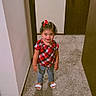 child, girl, smiling, standing, plaid_shirt, red_bow, curly_hair, sandals, carpet, hallway, door, wall, indoor, person, cute, happy, young_child, fashion, home, portrait