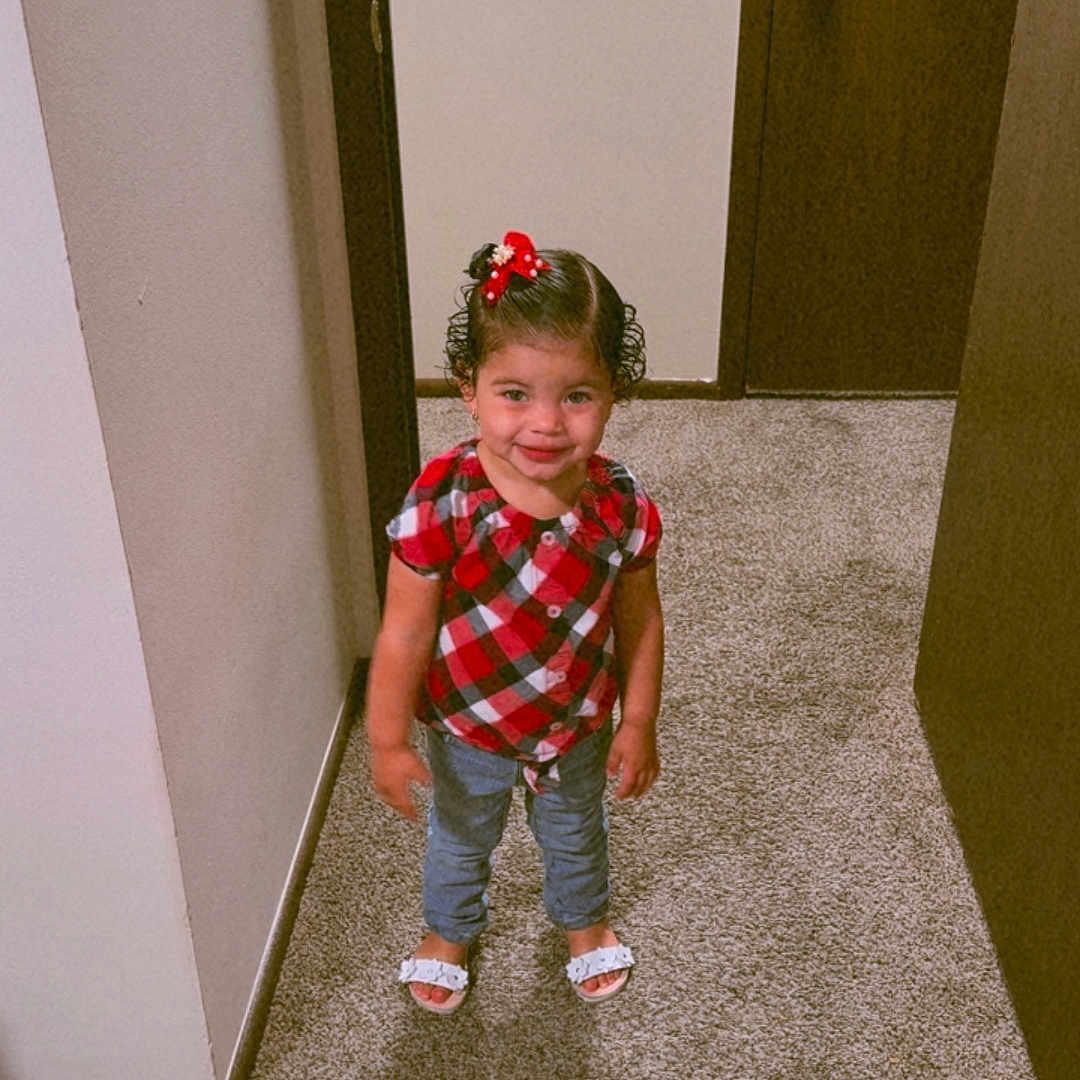 Isamar Victoria is registered to the contest to win money with this photo: child, girl, smiling, standing, plaid_shirt, red_bow, curly_hair, sandals, carpet, hallway, door, wall, indoor, person, cute, happy, young_child, fashion, home, portrait