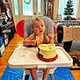 child, baby, high_chair, cake, birthday, frosting, messy, indoor, home, wood_floor, balloon, table, window, curtains, carpet, cabinet, picture_frame, christmas_tree, footrest, face