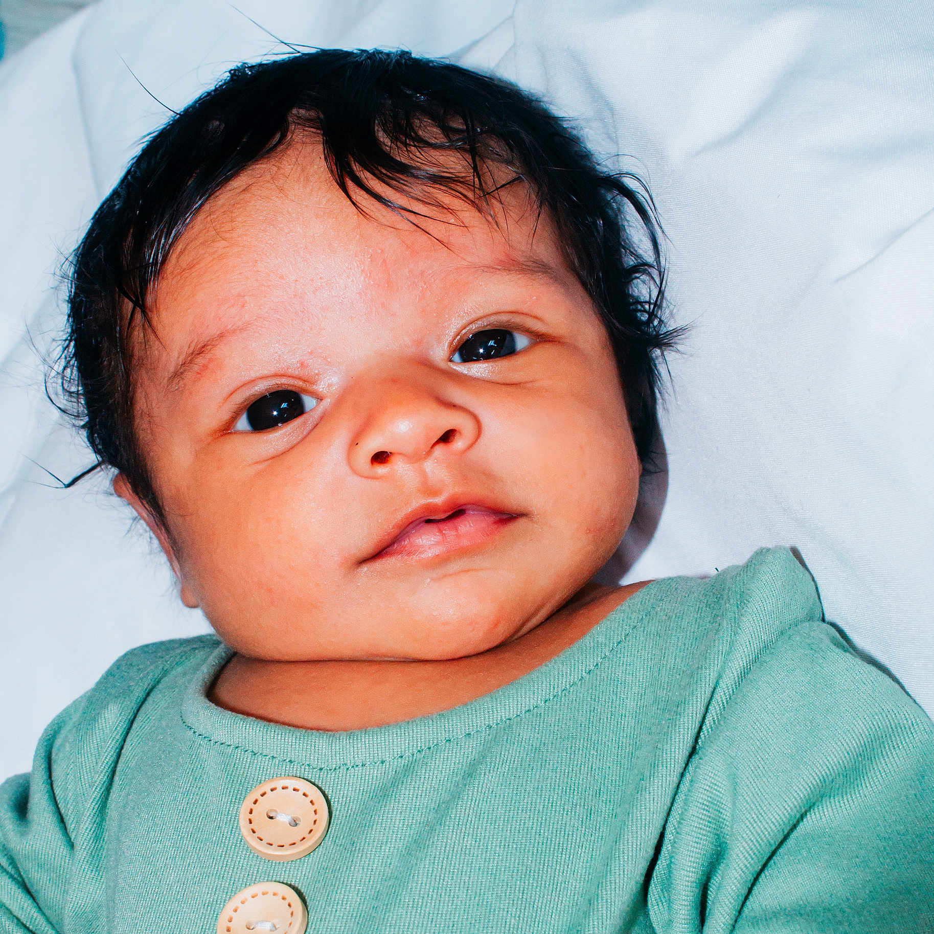 Zamir is registered to the contest to win money with this photo: baby, black_hair, child, close_up, cute, expression, eyes, face, green_clothing, indoors, infant, lying_down, newborn, person, portrait, skin, soft_light, white_background, wooden_buttons, young