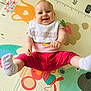 baby, child, smiling, bib, pink_pants, socks, play_mat, colorful, floor, happy, indoors, cute, infant, legs_up, joyful, person, toddler, patterned_mat, sitting, cheerful