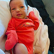 Faith is registered to the contest to win money with this photo: baby, newborn, infant, onesie, red_clothing, blanket, soft_texture, lying_down, cute, face, expression, hand, foot, indoors, person, child, sleepwear, portrait, resting, cozy