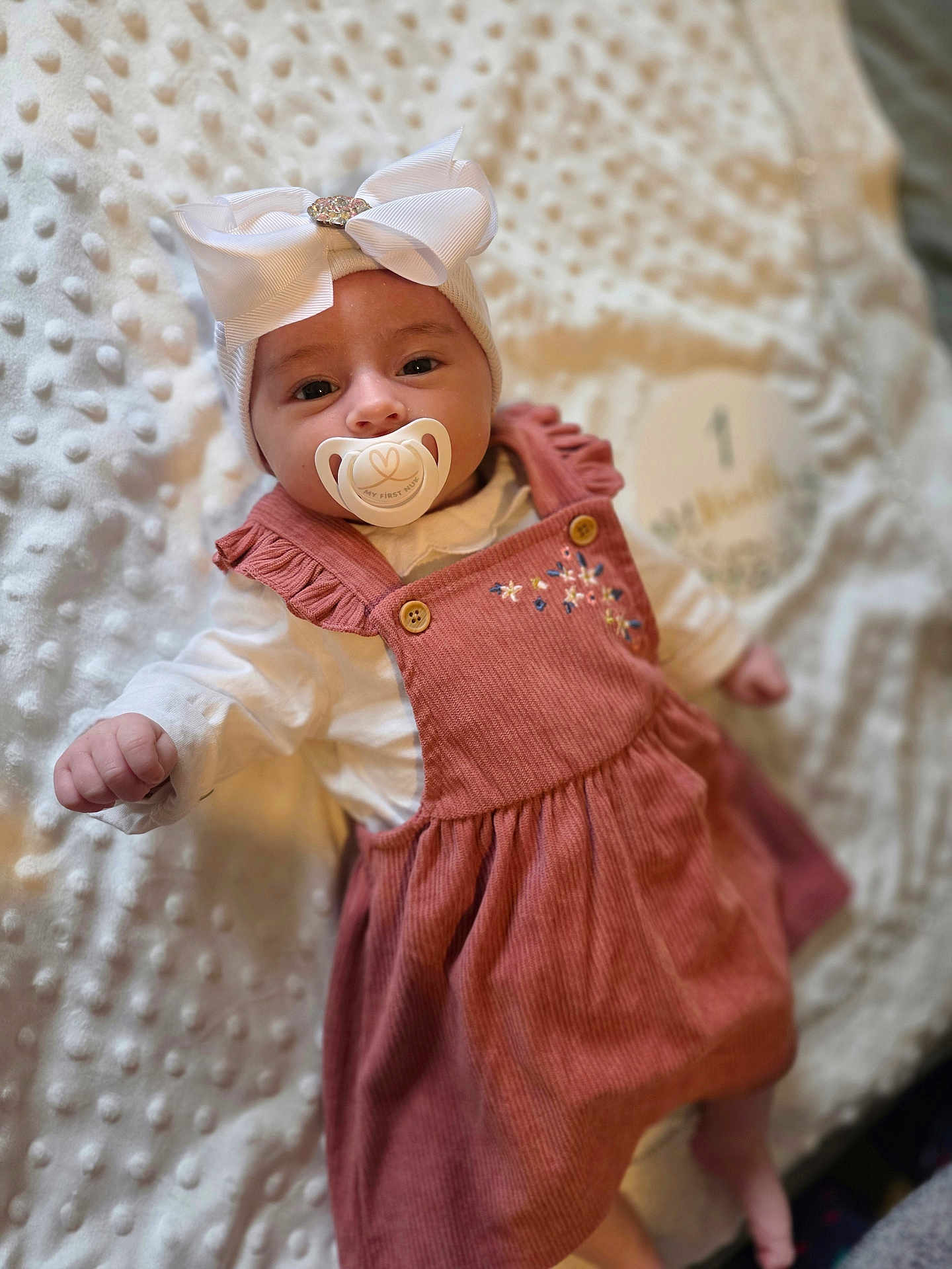 Maria participe au concours pour gagner de l'argent avec cette photo : baby, infant, pacifier, bow_headband, headband, blanket, textured_blanket, dress, corduroy_dress, pink_dress, white_bow, portrait, face, eyes_open, smile, milestone_disc, ruffle, long_sleeves, lying_down, soft_light