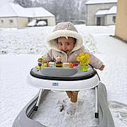 Thaïs participe au concours pour gagner de l'argent avec cette photo : baby, walker, snow, winter, outdoor, child, hooded_coat, toy, playful, cute, cold, footwear, snowfall, face, person, infant, background, house, daylight, smile