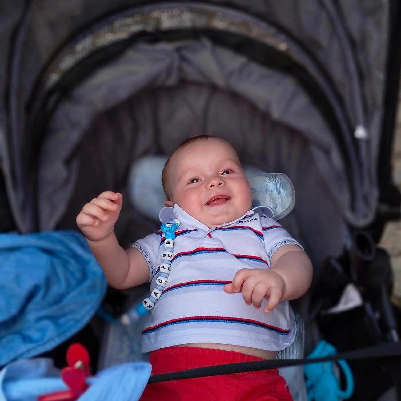 Kacper is registered to the contest to win money with this photo: baby, baby_accessories, child, clothing, comfort, cute, fabric, happy, infant, outdoor, pacifier_clip, person, polo_shirt, portrait, red_shorts, relaxed, seat, smiling, stroller, young_child