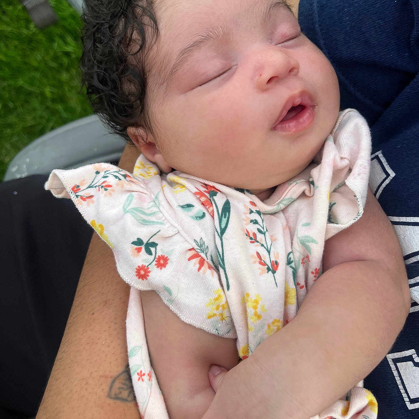 Evelyna is registered to the contest to win money with this photo: baby, sleeping, curly_hair, infant, arms, holding, floral_outfit, peaceful, close_up, person, skin, hand, resting, outdoor, greenery, nature, portrait, cute, child, toddler