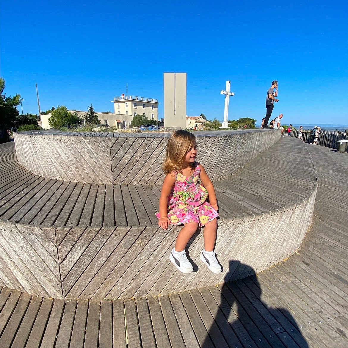 Lara participe au concours pour gagner de l'argent avec cette photo : architecture, bench, blue_sky, building, casual, child, cross, daytime, dress, girl, nature, outdoor, people, photographer_shadow, shadow, sneakers, sunny, tree, wooden_bench, wooden_floor