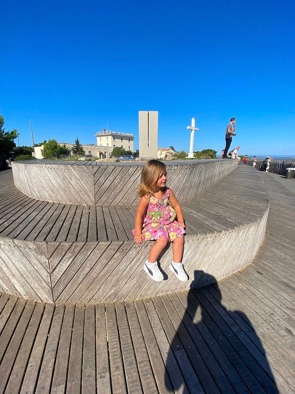 Lara participe au concours pour gagner de l'argent avec cette photo : architecture, bench, blue_sky, building, casual, child, cross, daytime, dress, girl, nature, outdoor, people, photographer_shadow, shadow, sneakers, sunny, tree, wooden_bench, wooden_floor