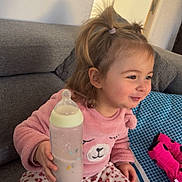 Lara participe au concours pour gagner de l'argent avec cette photo : baby_bottle, casual_clothing, child, childhood, couch, cozy, cute, happy, home, indoor, leopard_print_pants, person, pigtails, pillow, pink_sweater, plate, smiling, snack, toddler, toy