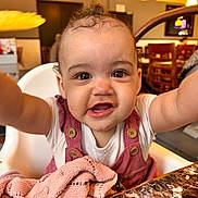 Kyra is registered to the contest to win money with this photo: baby, child, high_chair, pink_blanket, restaurant, table, happy, smiling, curly_hair, face, closeup, arms_outstretched, indoor, wooden_chair, buttoned_overall, white_shirt, marble_table, warm_lighting, person, cute