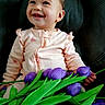 baby, child, smiling, pink_onesie, hearts, purple_tulips, flowers, green_leaves, sitting, black_leather, happy, portrait, cute, infant, closeup, indoor, person, flower_bouquet, natural_light, toddler